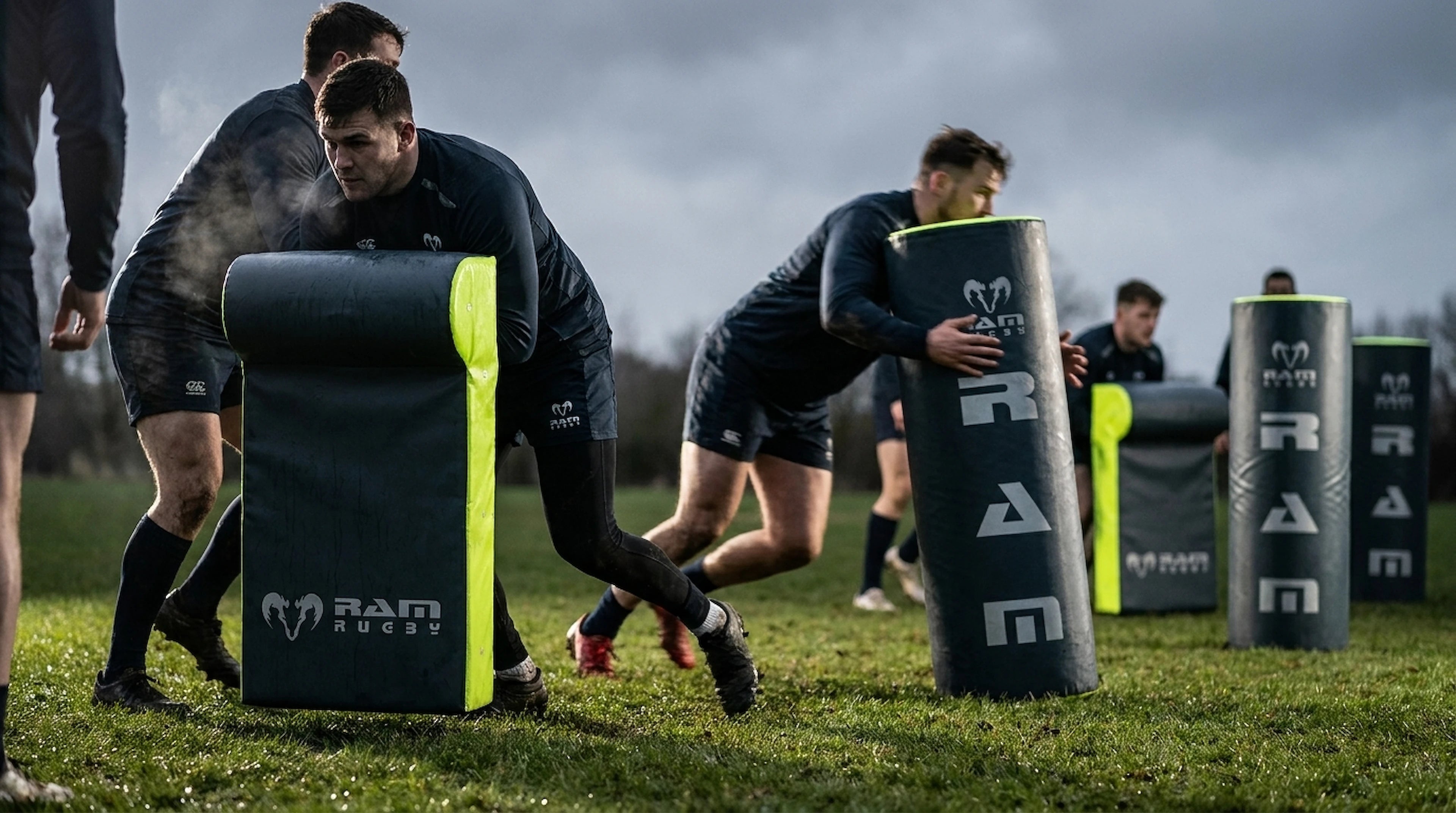 Athletes training with black and neon yellow rugby scrum pads on a grassy field under an overcast sky. The pads feature the RAM Rugby logo.